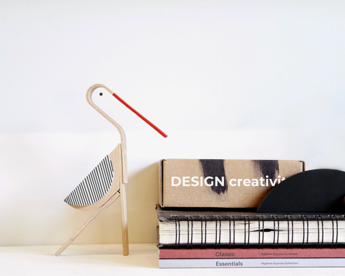 A modern wooden desk accessory shaped like a stork, featuring a light natural wood body, a red beak, and a striped wing, styled next to a stack of books and a box on a white surface against a white wall.