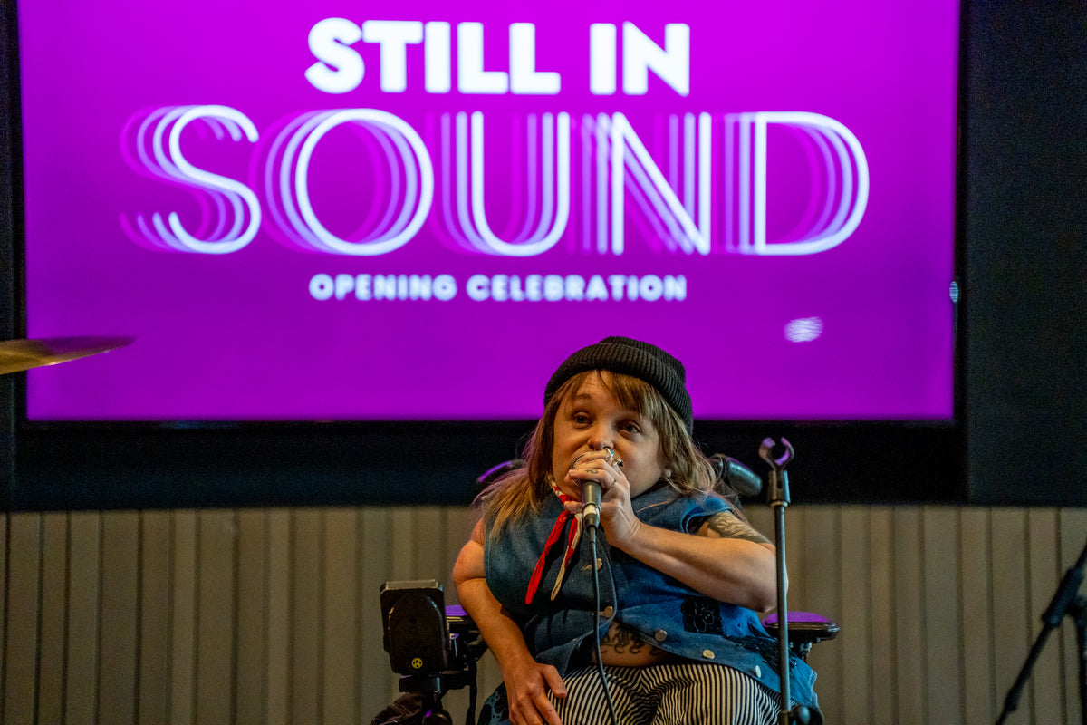 A person in a wheelchair singing into a microphone on stage, with a large purple screen behind them reading 'STILL IN SOUND OPENING CELEBRATION'.