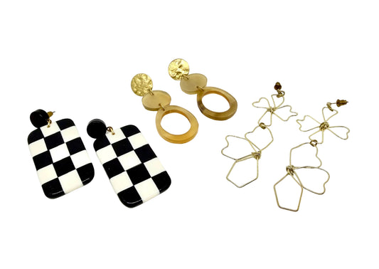 Three pairs of earrings on a white background, one black and white checkered, one brass and horn, the other gold flowers.