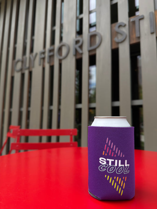 A purple can cooler sleeve with the words 'STILL COOL' and colorful diagonal line graphics, holding a silver can on a bright red table in front of a modern building with vertical slats and the word 'CLYFFORD' blurred in the background.