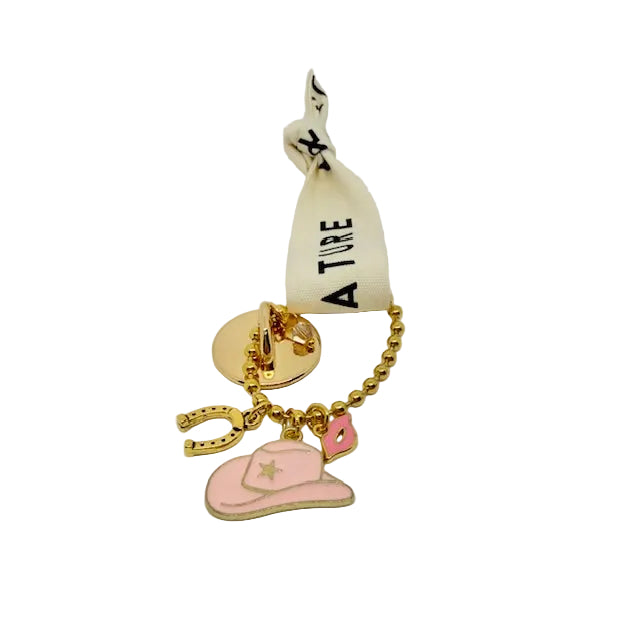 A gold-tone charm keychain featuring a pink cowboy hat, pink heart, gold horseshoe, and a pink star, attached to a gold ball chain and a cream ribbon with black text.