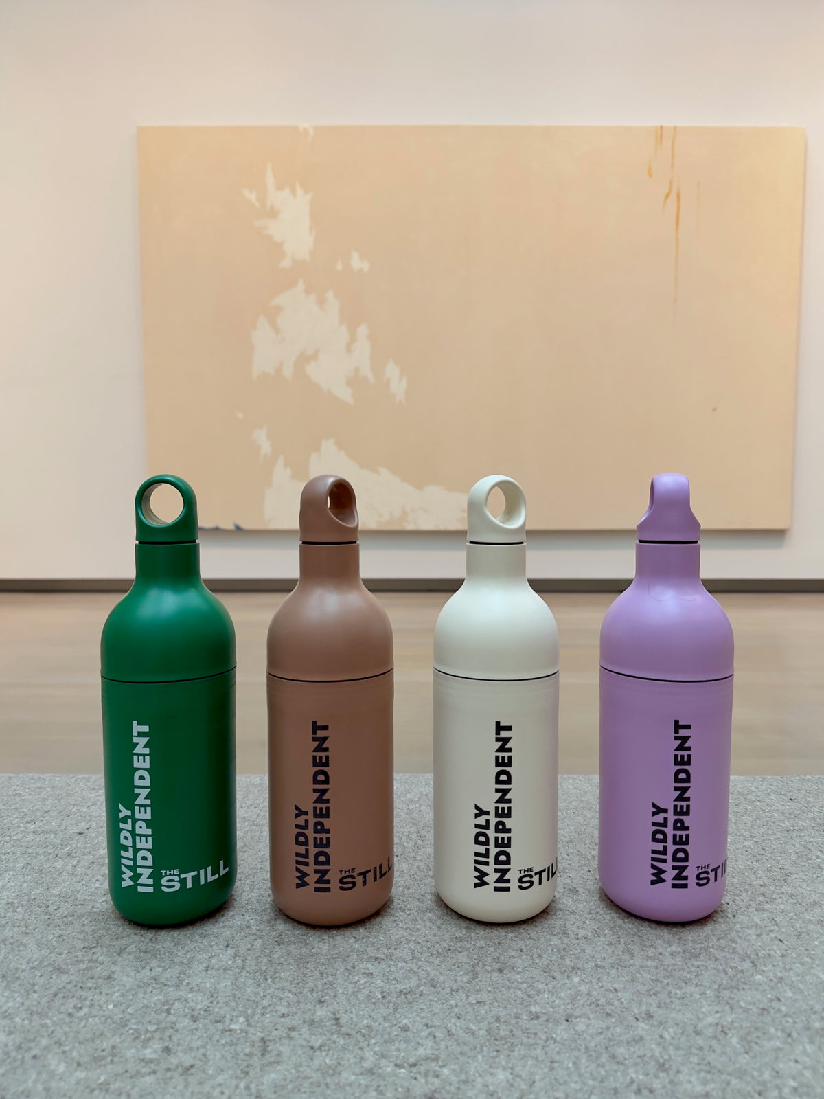 Four water bottles in green, brown, off-white, and lavender, all with the vertical text 'WILDLY INDEPENDENT STILL' on the front, arranged on a gray surface in front of a beige wall with a large abstract canvas.