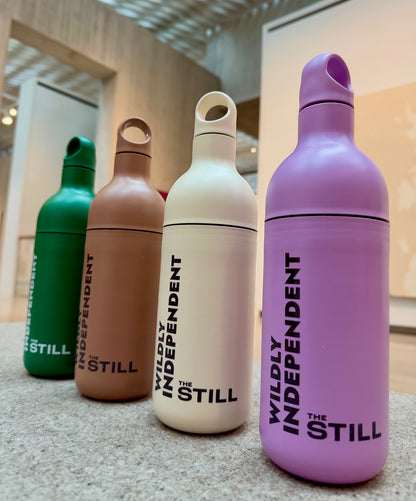 Four water bottle-shaped tumblers in green, tan, cream, and lavender, all printed with the vertical text 'WILDLY INDEPENDENT THE STILL', arranged on a countertop in an indoor setting.