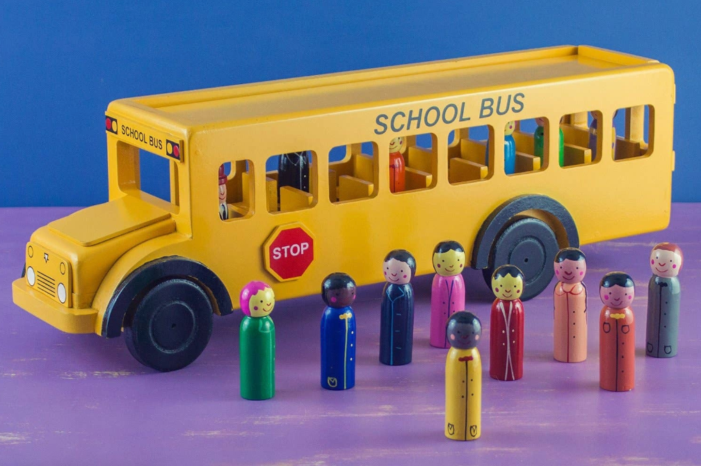 A handmade wooden school bus toy with wooden children figures in front, set against a blue and purple background.