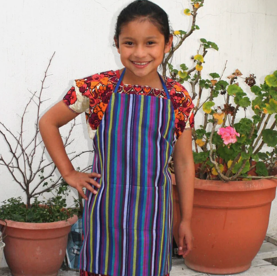 Young girl wearing a colorful striped apron standing in front of potted plants.