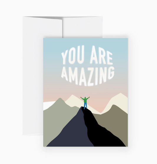 A greeting card with a white envelope behind it, featuring the text 'YOU ARE AMAZING' above a minimalist mountain landscape with a small person standing on a peak.