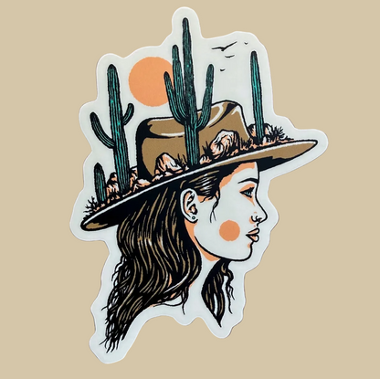 A die-cut vinyl sticker featuring a stylized desert illustration of a woman in profile wearing a wide-brimmed hat with cacti, photographed on a wooden surface.