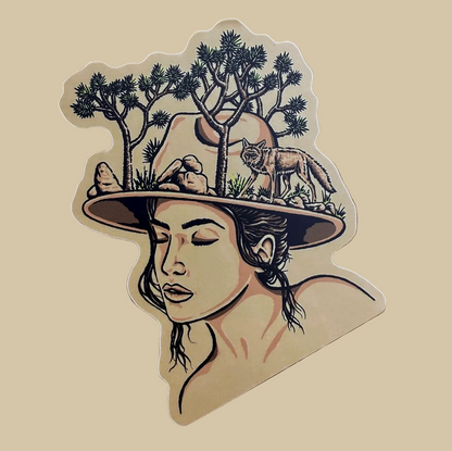 A die-cut sticker featuring an illustrated portrait of a woman wearing a wide-brimmed hat with desert trees, a deer, and a wolf, placed on a wooden surface.