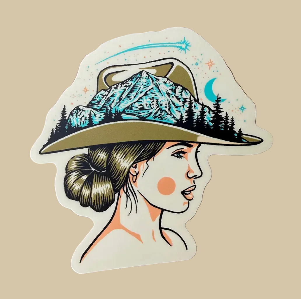 A die-cut vinyl sticker featuring an illustration of a woman in profile wearing a wide-brimmed hat with a mountain and pine forest design, placed on a wooden surface.