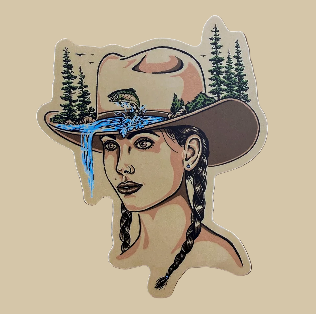 A die-cut vinyl sticker featuring an illustrated woman in a cowboy hat with pine trees, a fish, and blue water details, placed on a wooden surface.
