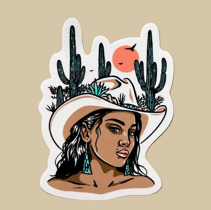 A die-cut vinyl sticker featuring an illustrated woman in a cowboy hat with cacti and a sun, placed on a textured tan paper background.