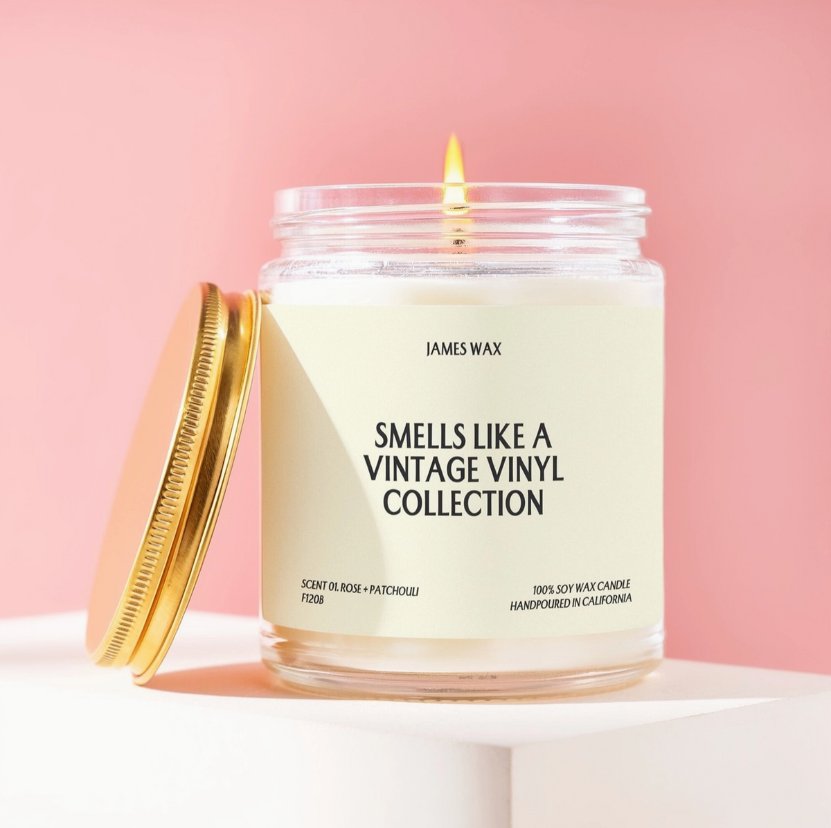 A lit James Wax candle in a clear glass jar with a cream label reading 'SMELLS LIKE A VINTAGE VINYL COLLECTION,' shown with a gold screw-top lid leaning beside it on a white pedestal against a soft pink background.