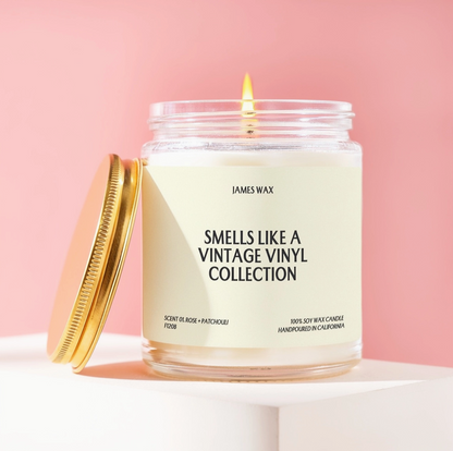 A lit James Wax candle in a clear glass jar with a cream label reading 'SMELLS LIKE A VINTAGE VINYL COLLECTION,' shown with a gold screw-top lid leaning beside it on a white pedestal against a soft pink background.