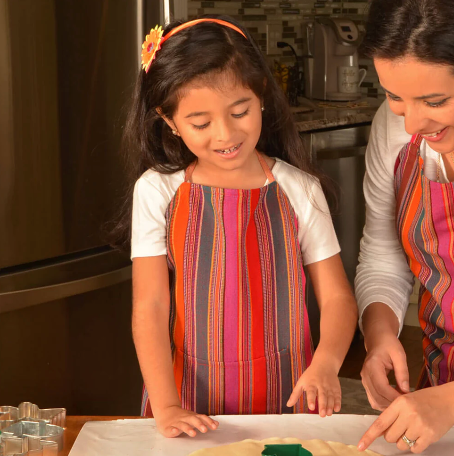 Woman and young girl in a kitchen, both wearing colorful aprons, engaged in baking.