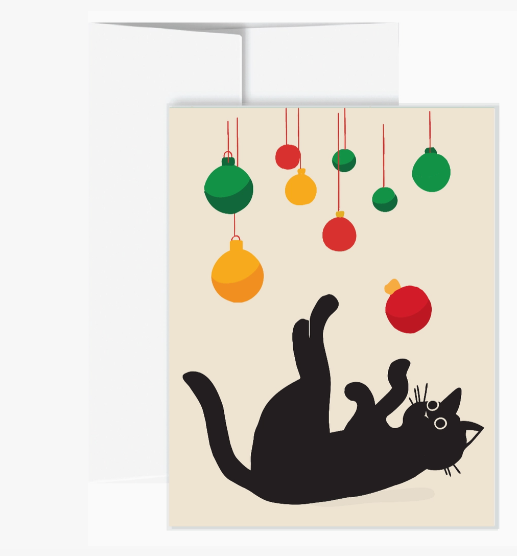 A greeting card featuring a black cat rolling on its back under hanging Christmas ornaments in green, red, and yellow, with a cream envelope behind it on a white background.