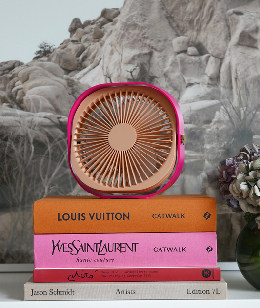 A small pink and beige portable fan sits on top of a stack of four fashion books, including Louis Vuitton and Yves Saint Laurent, with a gray rock wall backdrop and a flower vase at the side.