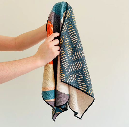 A person's hands holding up a square athletic towel with a geometric pattern. The towel features a dark teal blue-green side with beige diagonal line patterns, a cream side, and a side with orange, teal, and purple blocks.