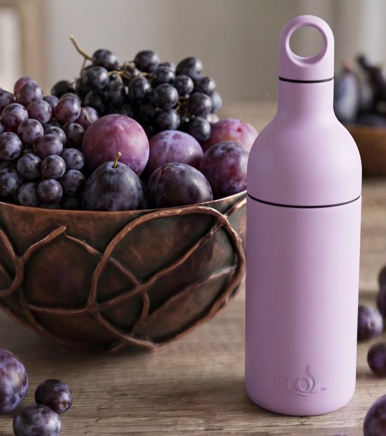A light purple bottle with a loop handle and black bands stands on a wooden table next to a copper bowl filled with purple grapes and plums.
