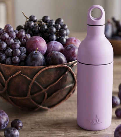 A light purple bottle with a loop handle and black bands stands on a wooden table next to a copper bowl filled with purple grapes and plums.