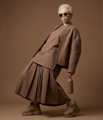 A model with short blonde hair and sunglasses poses in a matching brown oversized jacket, pleated skirt, and chunky sneakers against a solid brown studio background, holding a brown bottle.