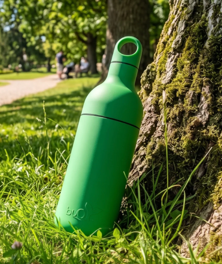 A bright green reusable bottle with a loop handle and a small embossed logo near the base, leaning against a mossy tree in a sunlit park.
