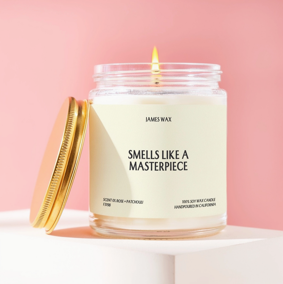 A lit James Wax candle in a clear glass jar with a cream-colored label and a gold-toned lid leaning beside it, set on a white pedestal against a soft pink background.