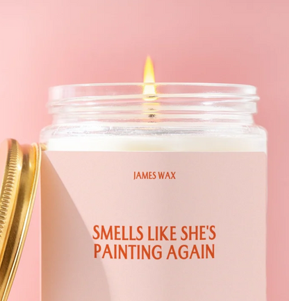 A lit candle in a clear glass jar with a pale pink label reading 'JAMES WAX' and 'SMELLS LIKE SHE'S PAINTING AGAIN' in orange text. A gold lid is partially visible on the left against a soft pink background.