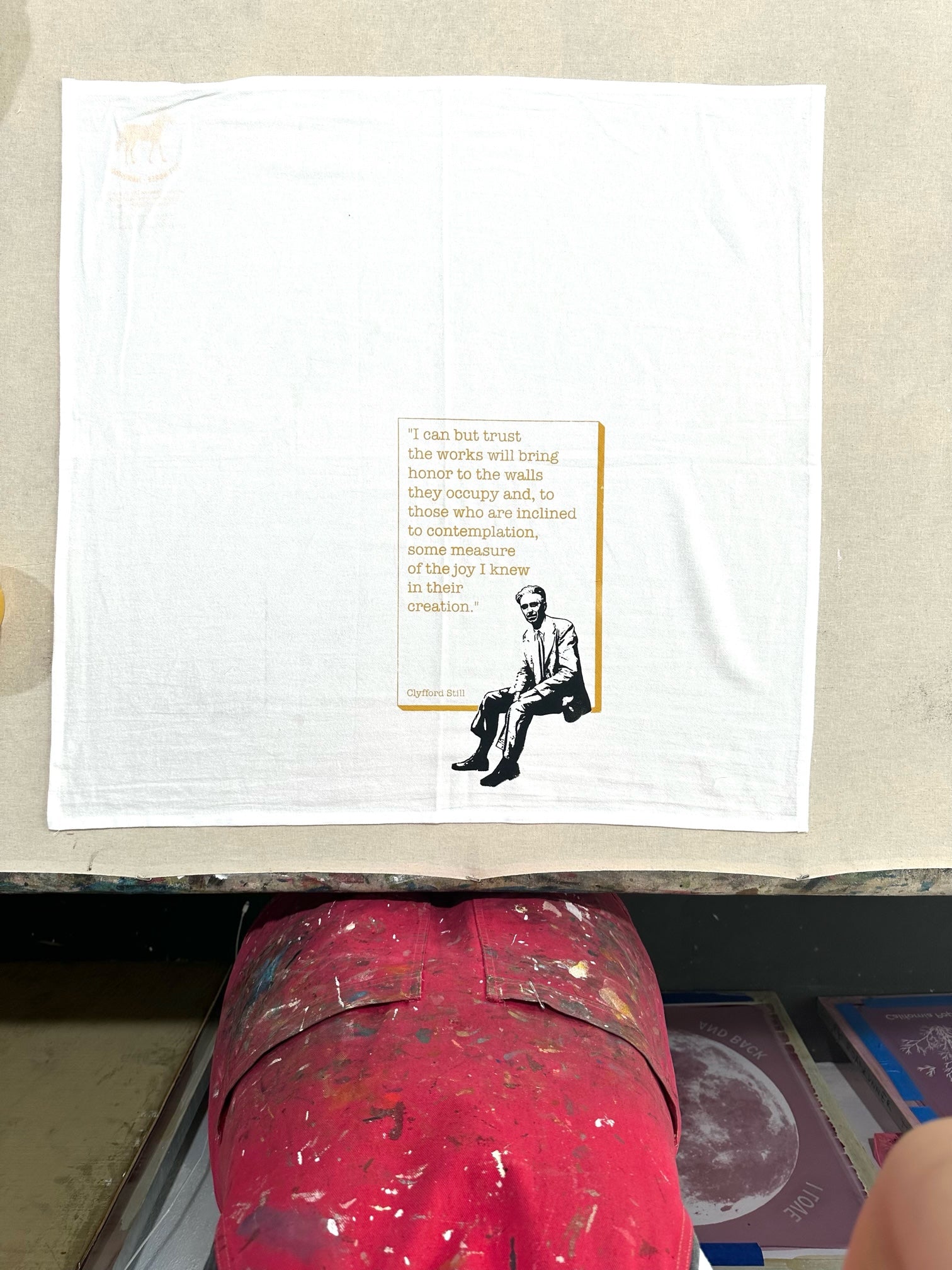 Top view of a white dish towel with the quote “ I can but trust the works will bring honor to the walls they occupy and, to those who are inclined to contemplation, some measure of the the joy I knew in their creation.” And a black and white graphic of Clyfford Still.