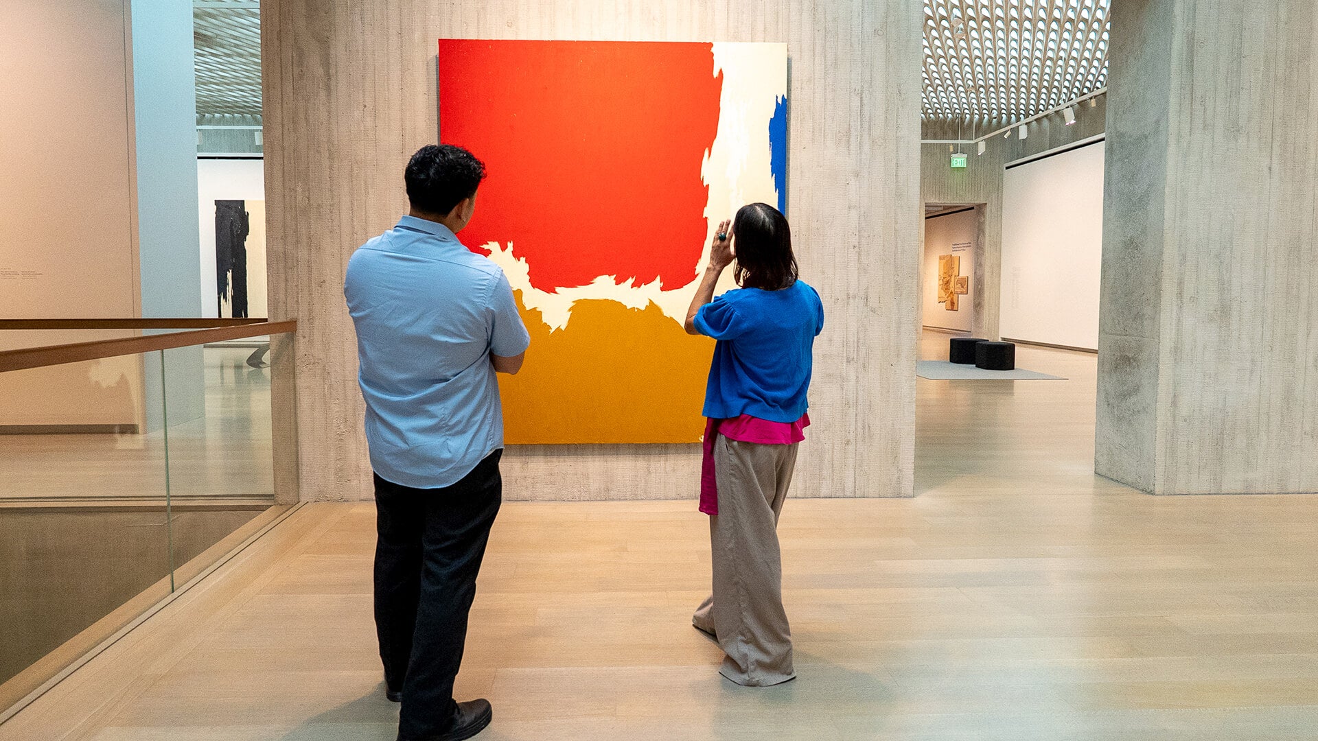 Two people in a museum looking at a large abstract painting with red, blue, white, and ochre sections.